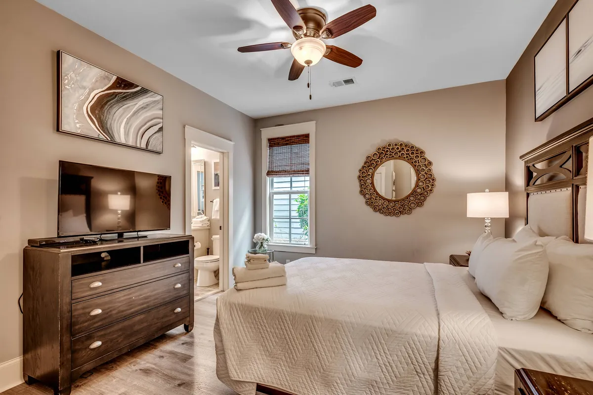 A serene bedroom featuring a comfortable bed dressed in soft linens, a TV for personal entertainment, a decorative mirror, and warm natural light streaming in through the window. Tasteful wall art and an inviting atmosphere make this space ideal for relaxation and privacy.
