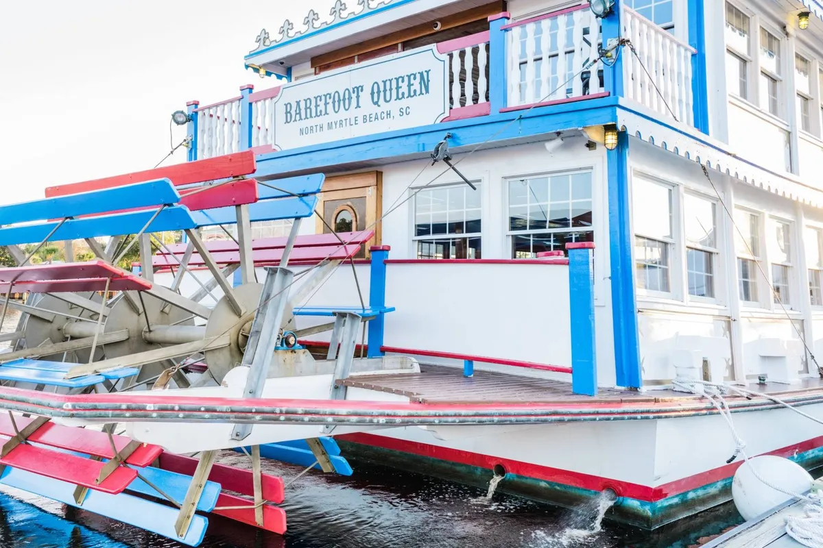 A colorful paddlewheel boat named 'Barefoot Queen' docked beside the water, with signage indicating sightseeing cruises. The festive exterior and scenic waterfront location evoke a sense of local adventure and fun.