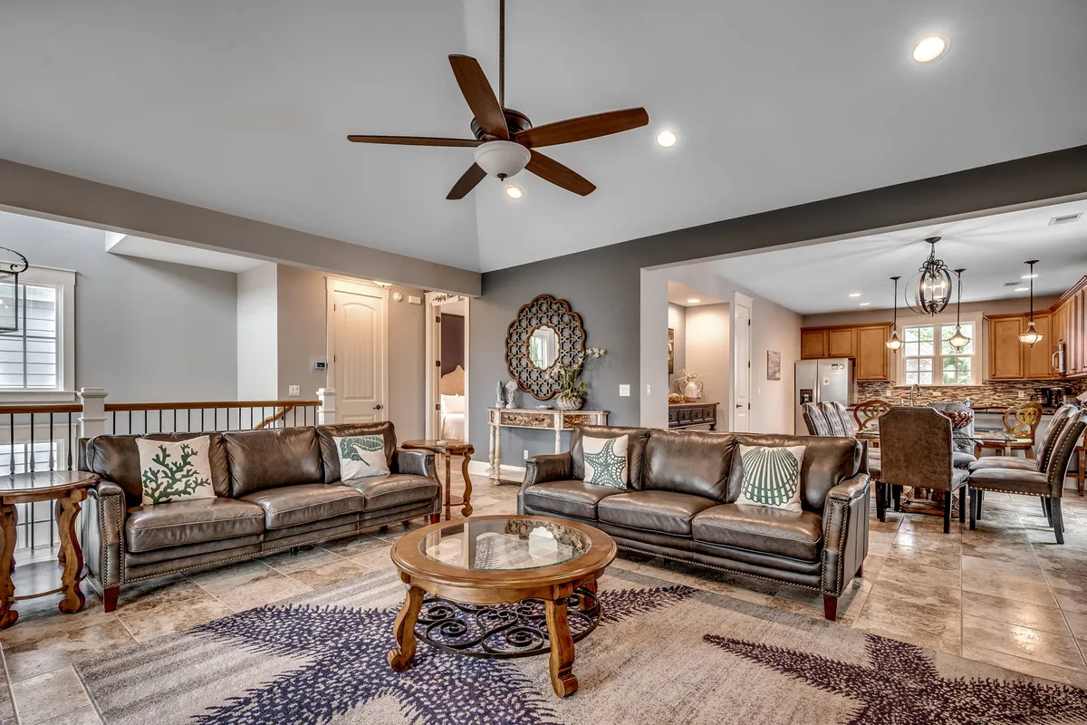 A cozy living room furnished with leather sofas, decorative pillows, a coffee table, and a view into the adjacent dining area. The space exudes comfort and is clearly suited for group activities, conversation, and relaxation.