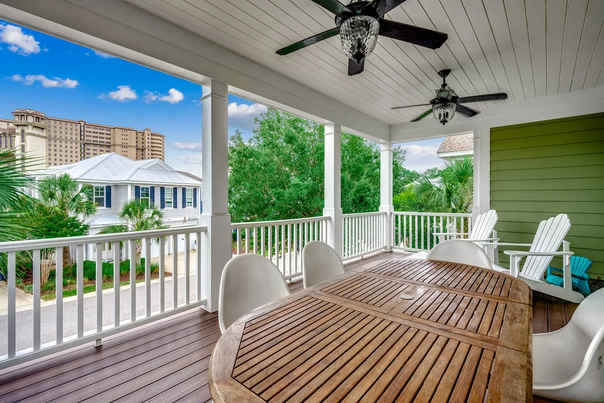 An exterior view of a covered outdoor dining area featuring a table, chairs, ceiling fans, lush green wall, and a glimpse of neighboring houses. The space invites outdoor relaxation and social gatherings.