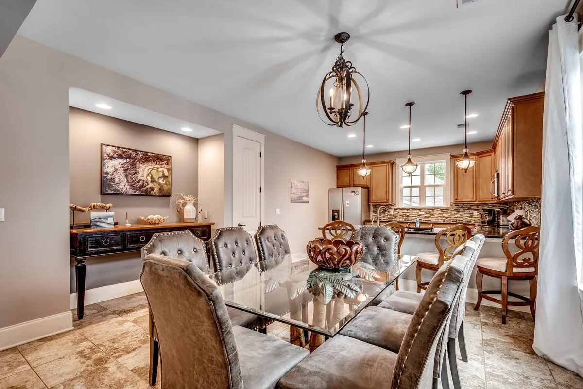 A dining area featuring a large table set beneath a chandelier, with kitchen cabinets and colorful artwork adding warmth and character. The space is clearly designed for family meals and entertaining guests.