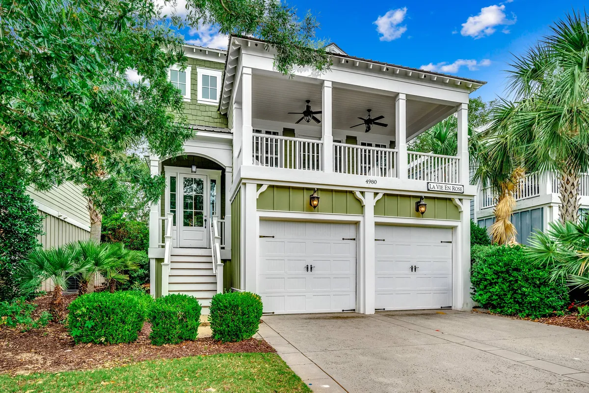 The image showcases the front exterior of a charming single-family house in North Myrtle Beach, with inviting green siding, a spacious porch complete with ceiling fans, welcoming front steps, and a beautifully landscaped garden. The porch exudes Southern hospitality, perfect for relaxing or gathering with family while enjoying the lush surroundings.