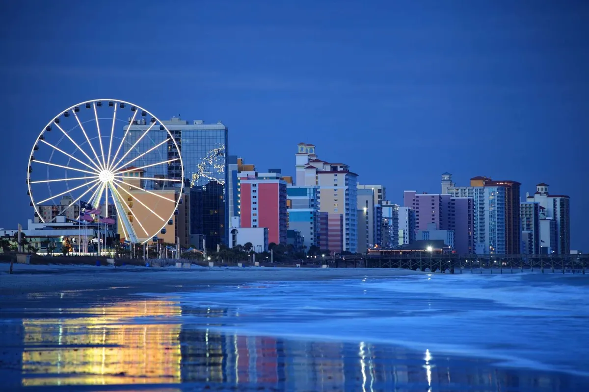 A lively view of a beachfront amusement area with a Ferris wheel, colorful buildings, and the ocean reflecting the evening lights—capturing the energy and entertainment of the nearby attractions.