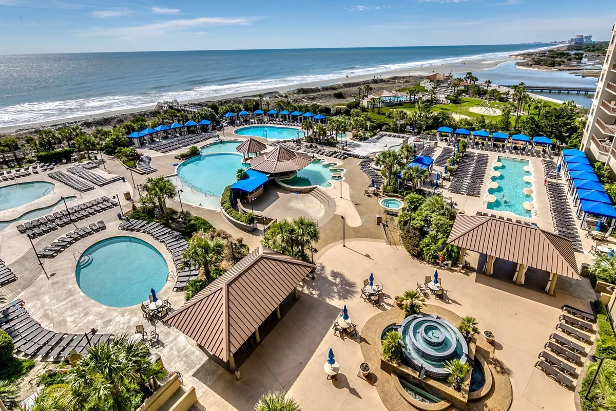 A vibrant exterior pool complex scene featuring multiple swimming pools surrounded by palm trees, lounge chairs, cabana structures, and an ocean view in the distance. The atmosphere is lively, with people swimming, sunbathing, and socializing.