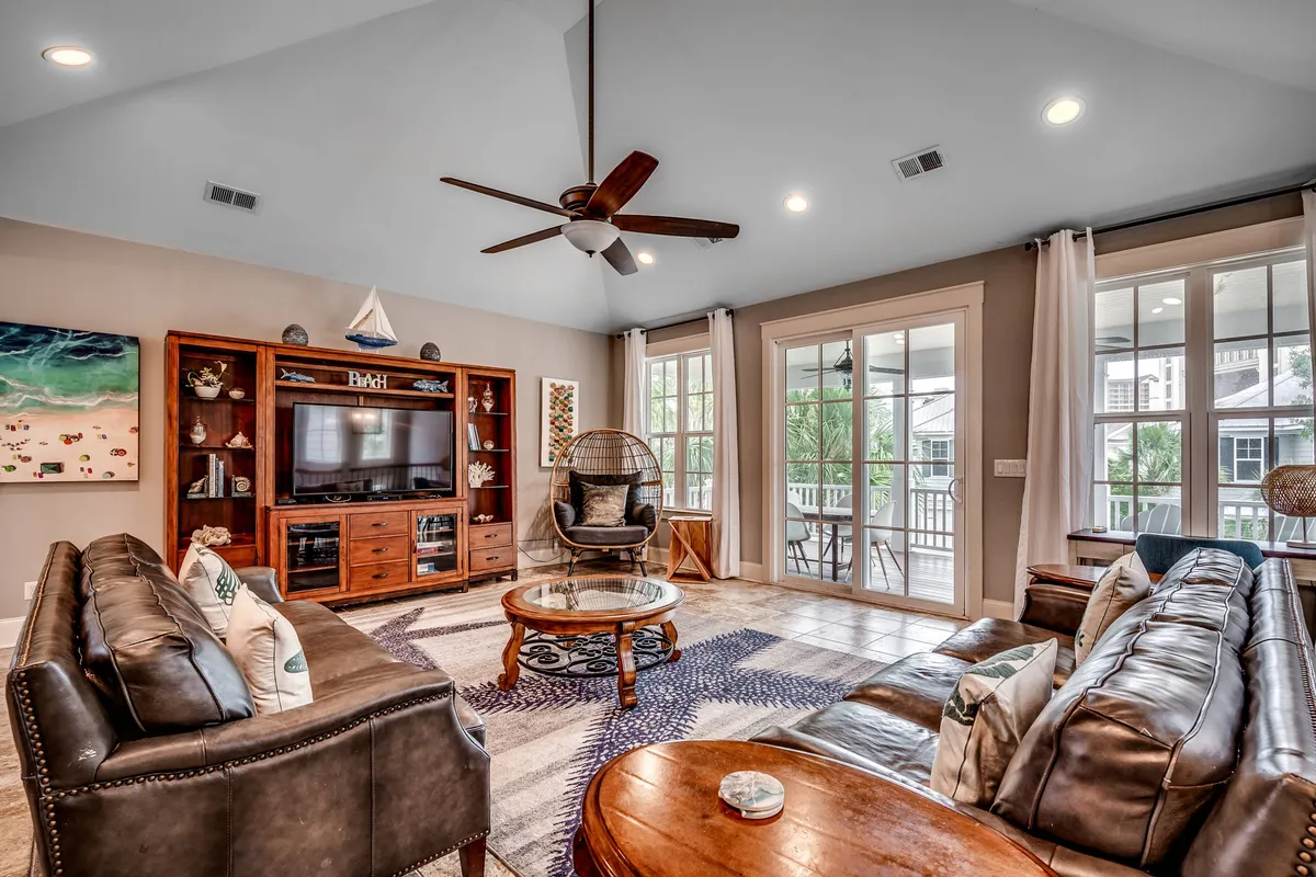 A cozy living space with large windows allowing natural light, comfortable seating options including a hanging chair, and a wooden entertainment center. The setup encourages relaxation, socializing, and enjoying downtime between beach outings or visits to Barefoot Landing.