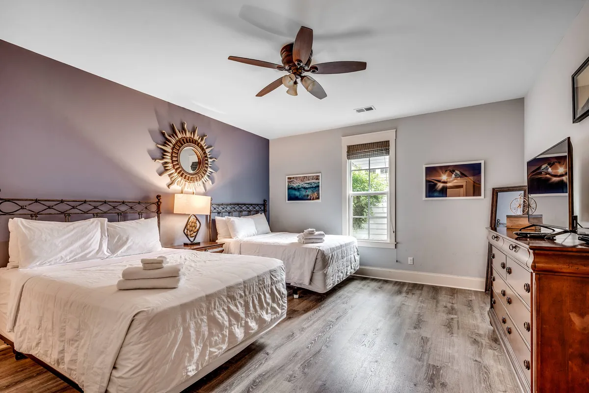 A bright, welcoming guest room with two beds featuring crisp white linens, a decorative mirror, a wooden dresser, and calming artwork. The arrangement is perfect for families or friends sharing a space while still maintaining individual comfort.