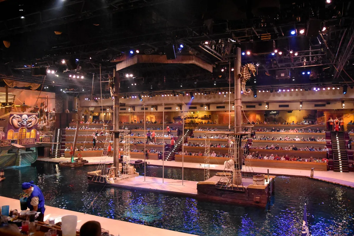 An immersive interior scene of a live performance venue featuring a large ship on stage surrounded by water, with audience seating visible—evoking the excitement of dinner shows like Pirate's Voyage.