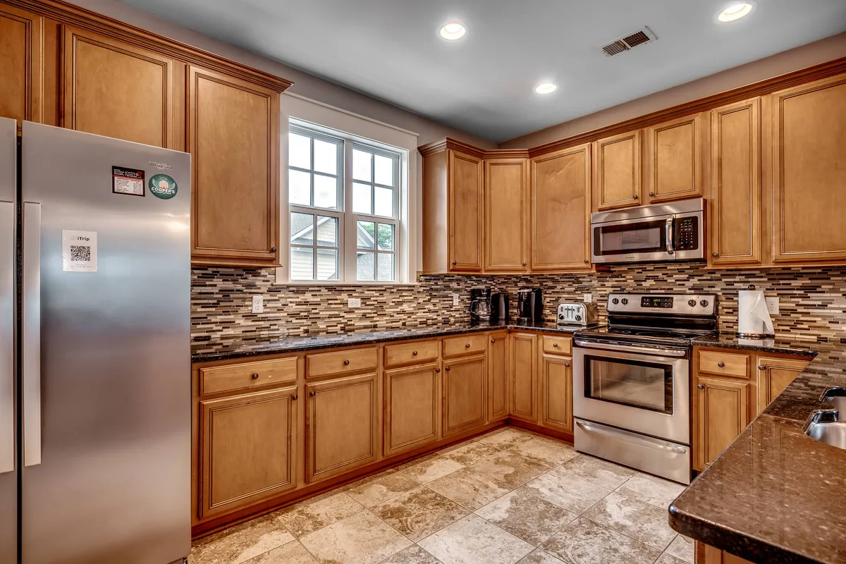 A modern kitchen with stainless steel appliances, wooden cabinetry, a black countertop, and a sunlit window. The space is practical and stylish, ideal for preparing meals and gathering with loved ones.