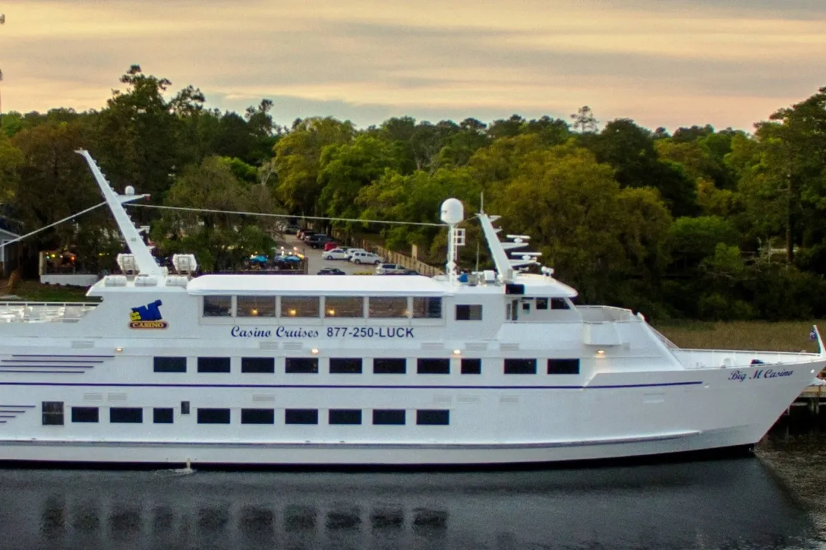 An exterior view of a large white yacht docked near lush greenery, with prominent casino signage visible. The scene is lively, with an ambiance that suggests entertainment, dining, and social experiences on the water.