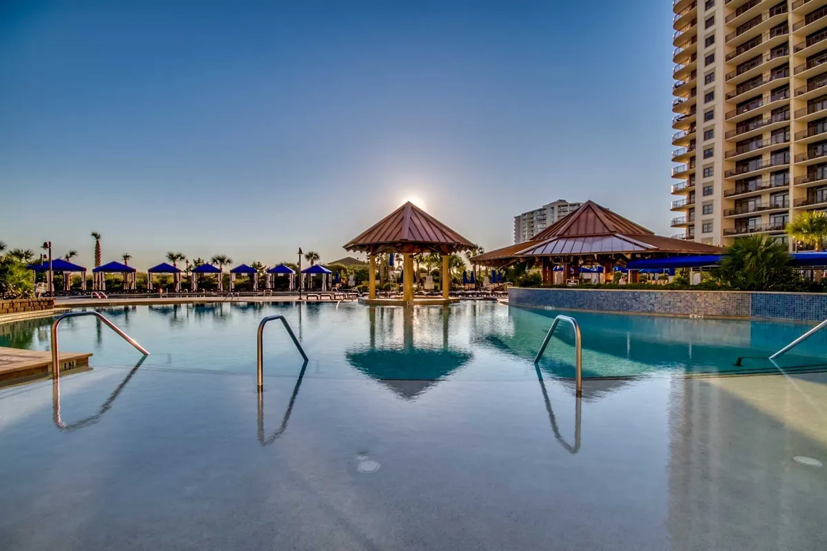A stunning exterior shot of the resort's swimming pool area, with sparkling blue water, poolside loungers, and shaded pavilions set against a backdrop of the main hotel building and a beautiful sunset sky.
