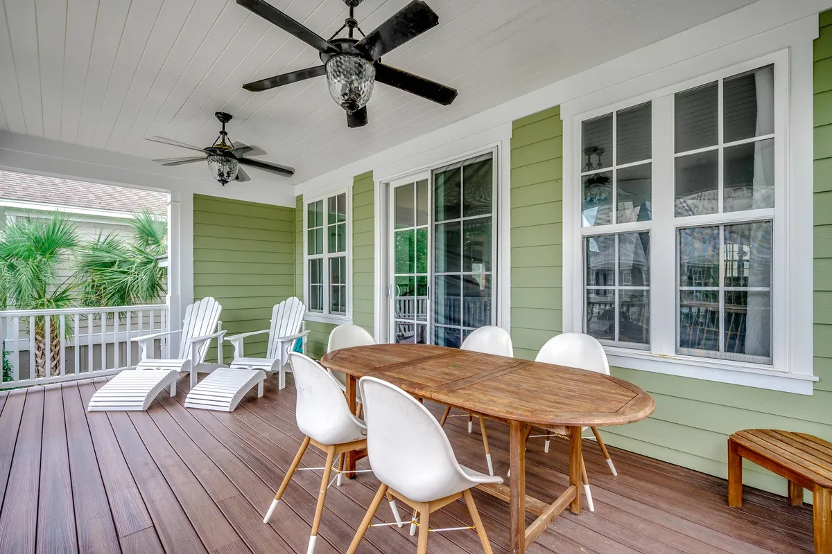A covered balcony with a wooden dining table, white chairs, Adirondack chairs, and ceiling fans, set against large windows that let in plenty of natural light. The space invites outdoor dining and relaxed conversations in a breezy, comfortable setting.