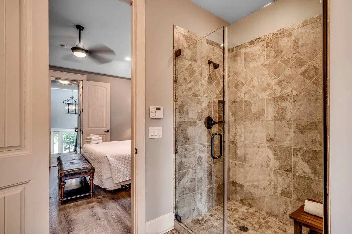 A modern ensuite bathroom with a glass shower enclosure, textured stone tiles, and a wooden bench. There is a pleasing view into a bedroom, emphasizing the connection between private sleeping quarters and luxurious, accessible bath spaces.